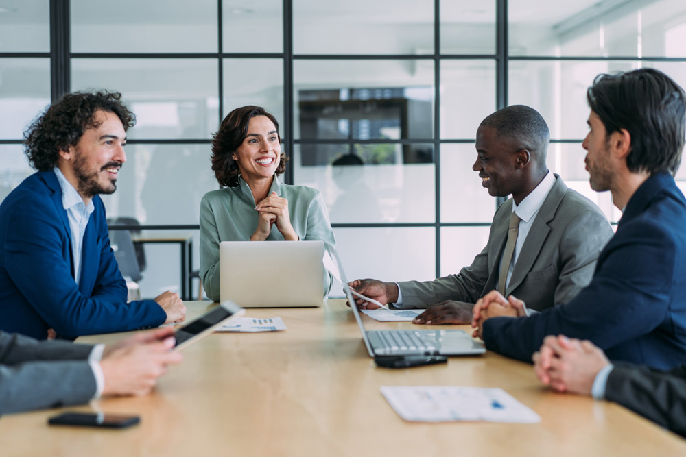 En grupp affärspersoner med multietnisk bakgrund sitter runt ett konferensbord i ett modernt styrelserum och håller ett professionellt affärsmöte.